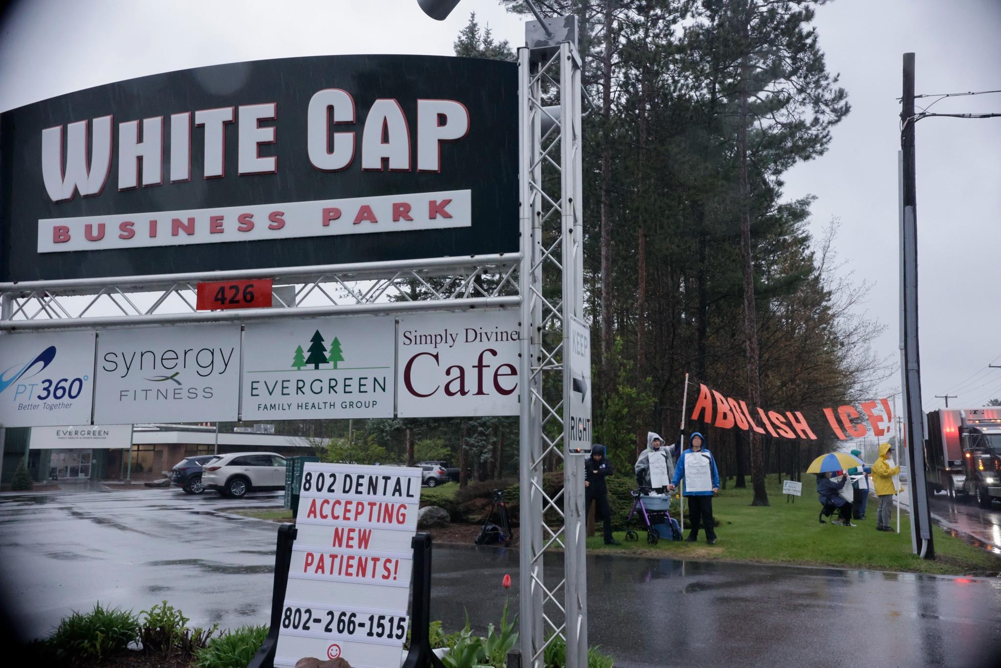 Photo of the entrance of White Cap Business Park, with people protesting in the background holding a sign that reads "Abolish ICE!"